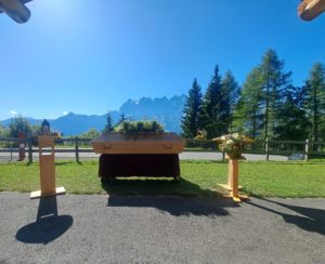 Cérémonie funéraire laïque en plein air, avec un cercueil en bois au centre, entouré de compositions florales sur des pupitres en bois. En arrière-plan, un paysage alpin majestueux sous un ciel bleu, avec des sapins et les montagnes des Dents du Midi, apportant une atmosphère paisible et solennelle.