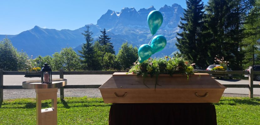 Cérémonie funéraire laïque en plein air, avec un cercueil en bois au centre, entouré de compositions florales sur des pupitres en bois. En arrière-plan, un paysage alpin majestueux sous un ciel bleu, avec des sapins et les montagnes des Dents du Midi, apportant une atmosphère paisible et solennelle.