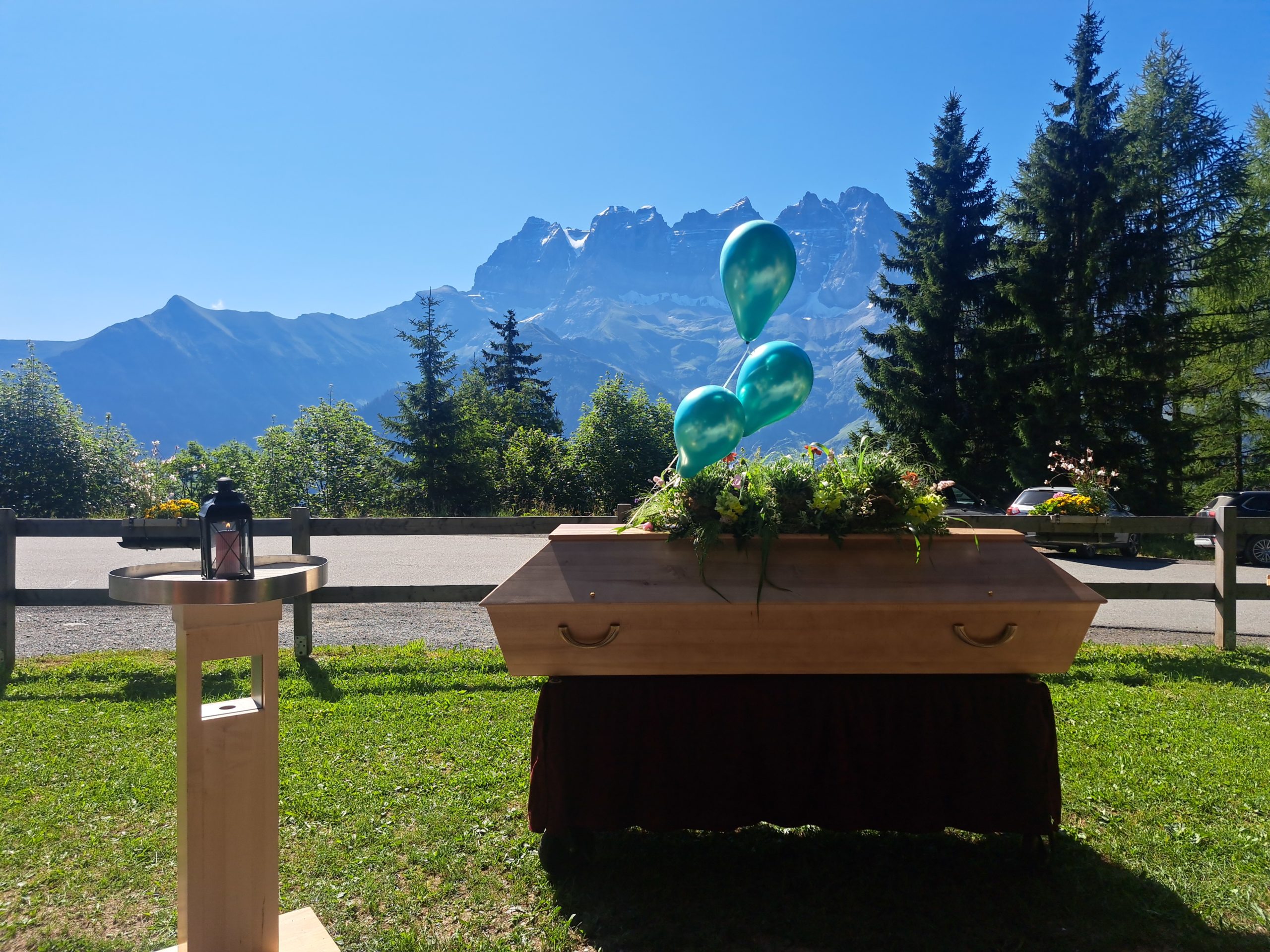 Cérémonie funéraire laïque en plein air, avec un cercueil en bois au centre, entouré de compositions florales sur des pupitres en bois. En arrière-plan, un paysage alpin majestueux sous un ciel bleu, avec des sapins et les montagnes des Dents du Midi, apportant une atmosphère paisible et solennelle.